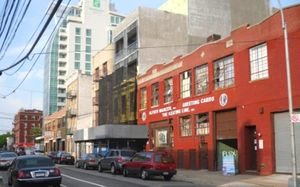 A red brick building beside a taller grey building. In the distance, a green and much taller, off-white colored building looms over the two. The street is lined with cars in front of the two buildings.