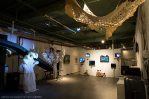 A gallery space with monitors lined up on the back wall with a white statue of a hand stands on the left. A large tree painted completely white stands next to the giant hand. A large, beige tapestry with cut outs hangs on the ceiling.