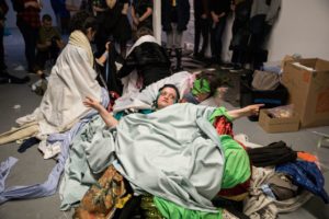 Four people covered in numerous blankets and clothing are rolling on the floor. A group of people stand by to take pictures of the scene. 