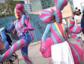Two people wearing morph suits that are bright pink and teal stripes are dancing in a parade as onlookers are nearby. One performer wears a white pant over their suit as another one adds a periwinkle blue to their suit.