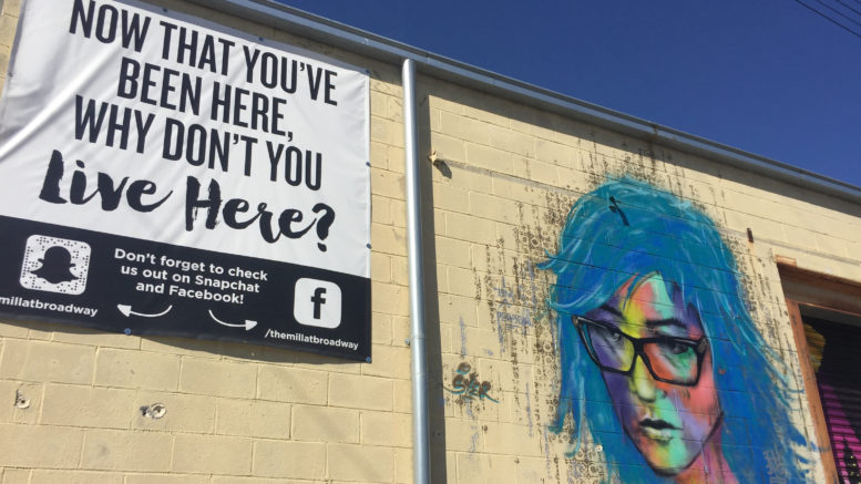 A sunny wall that looks like the side of a warehouse. Painted on the wall is an image of a woman with blue hair, glasses and a multicolored face. Next to the painting is a sign that says "Now that you've been here, why don't you live here?"