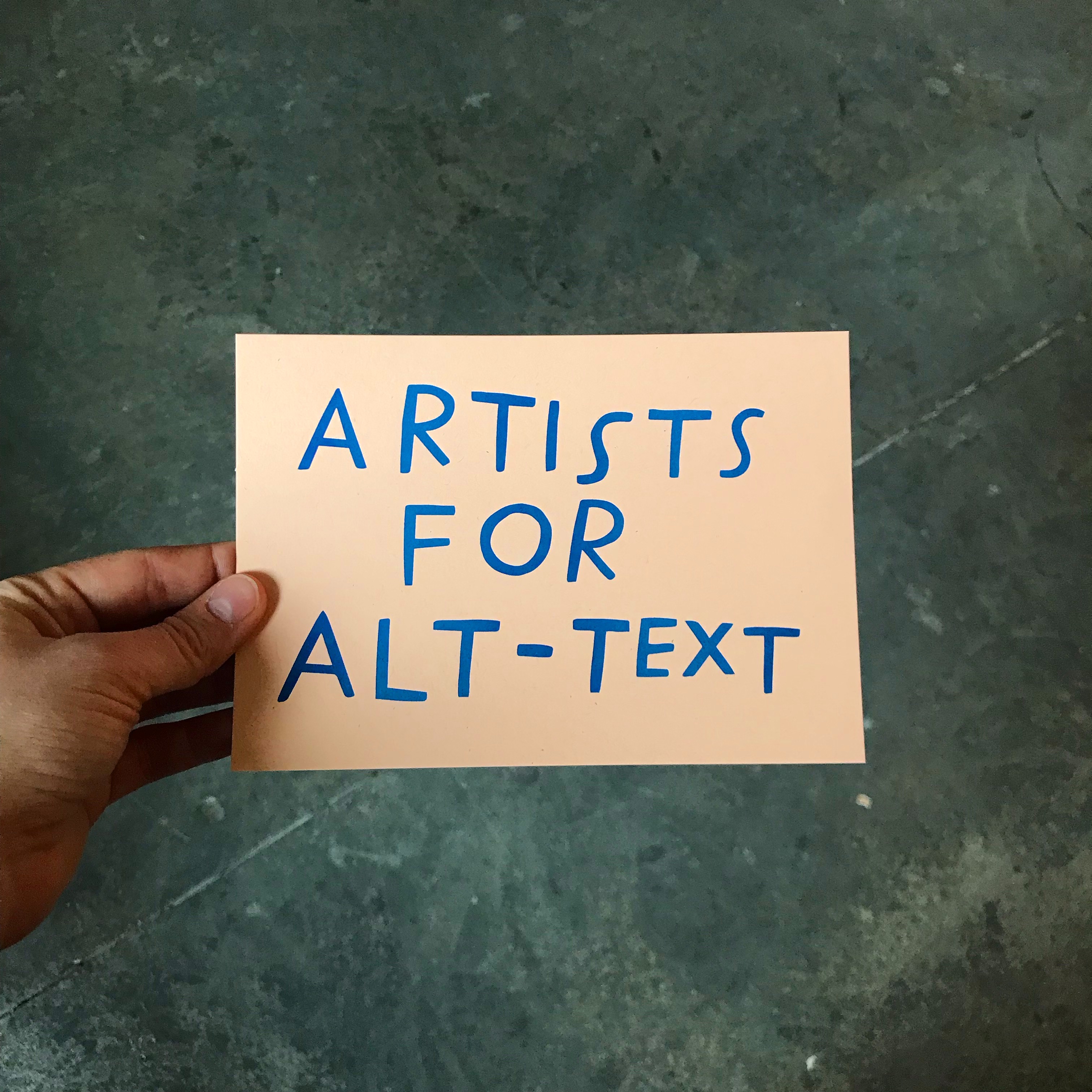 Image Description: A hand holds up a peach-coloured paper that reads “Artists for Alt-Text” in handwritten, dark blue letters. The background is a marbly, dark green floor.