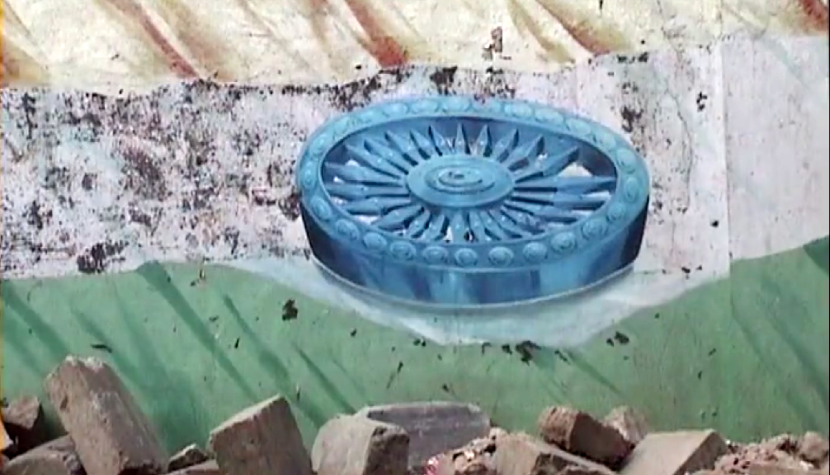 A decaying wall mural of the Indian flag, gold on the top a blue wheel in the middle and green on the bottom, with a pile of rubble and bricks on the bottom. 