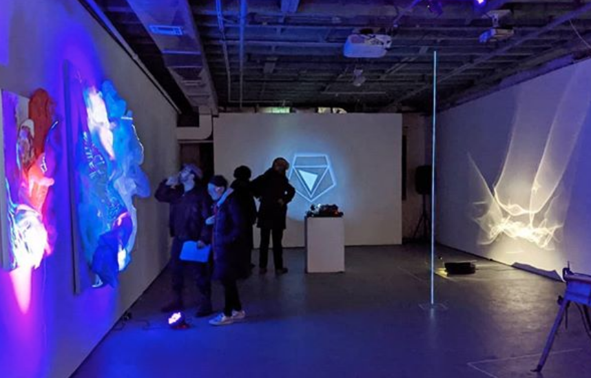 A darkened gallery with brightly colored light emanating from sculptures on the walls and floor. A small group of people looking at the artwork.