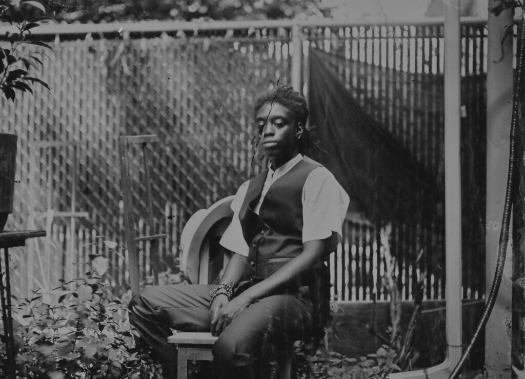 Black and white photo of a young person wearing a black suit vest, white short sleeve shirt and black dress pants sits on a chair in a backyard.