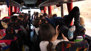 A photo taken from the back of children on a bus, one is lying flat on their back over the seats. 
