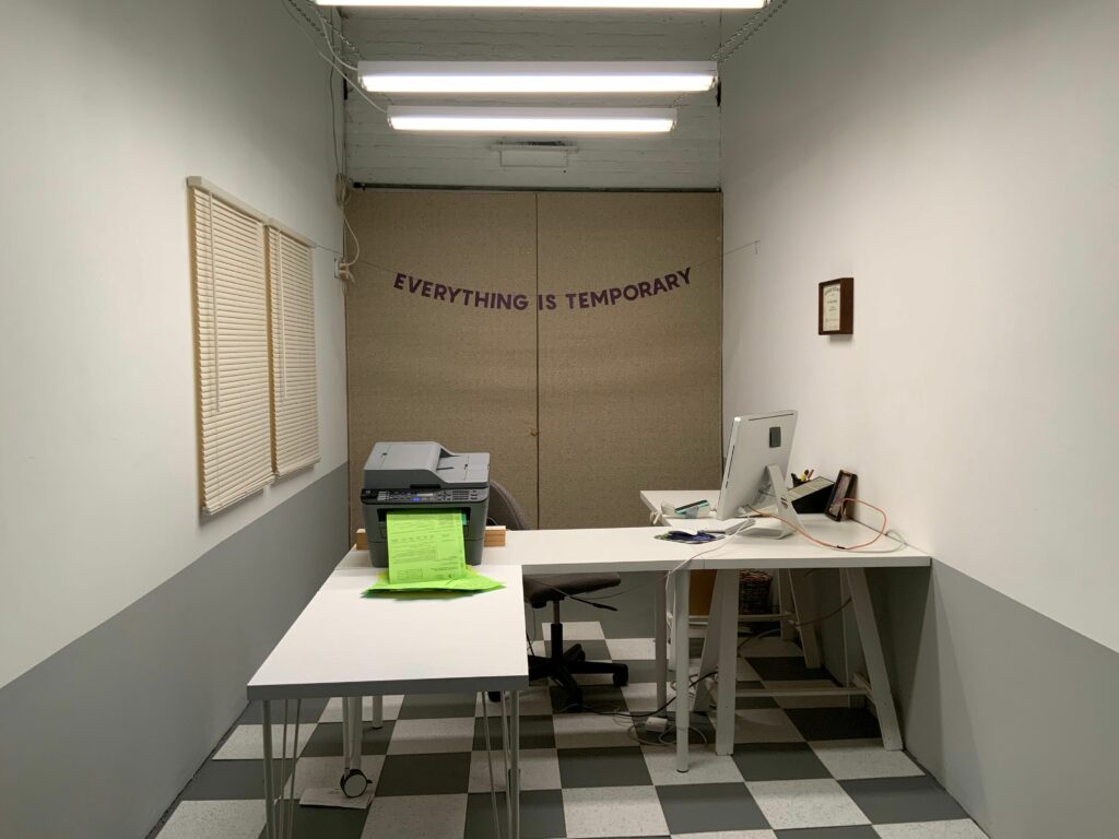 A small space set up with an L-shaped desk that holds a computer monitor, a printer with green paper, and stationary. The floor is grey and white checkered tile. The beige double doors behind the desk has a banner that says, "EVERYTHING IS TEMPORARY".