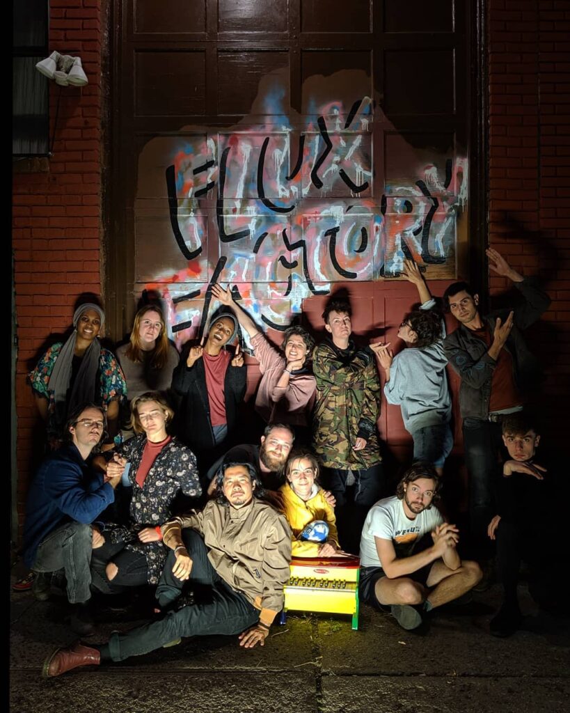 A group of Flux Factory Residents pose at night in front of the Flux Factory logo on the front of our building