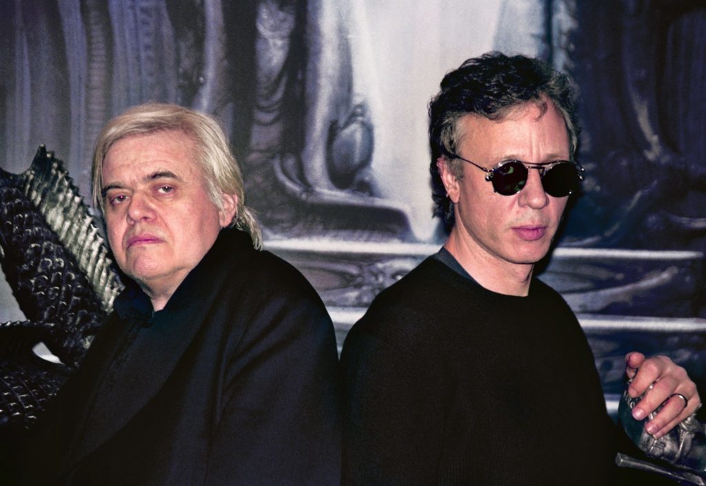 Image of Peter Gatien & HR Giger, dressed in all black and with their backs toward each other, posing.