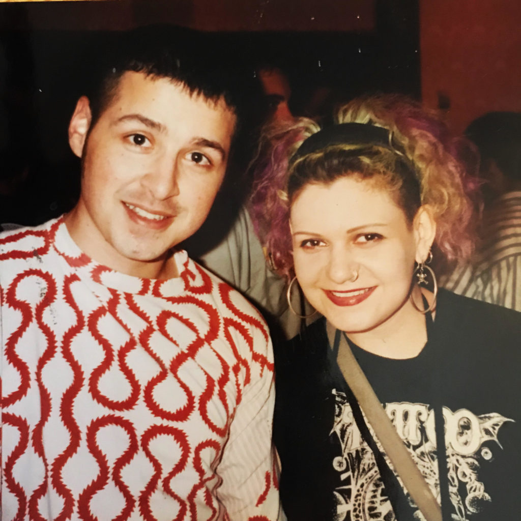 DJ Keoki & DJ Marlow standing next to each other, circa 1989. DJ Keoki is on the left with a white longsleeved shirt with red figure 8 patterns. DJ Marlow is on the right with a black shirt with a tattoo design that has white lettering in the center. The photo appears to be taken at a club.